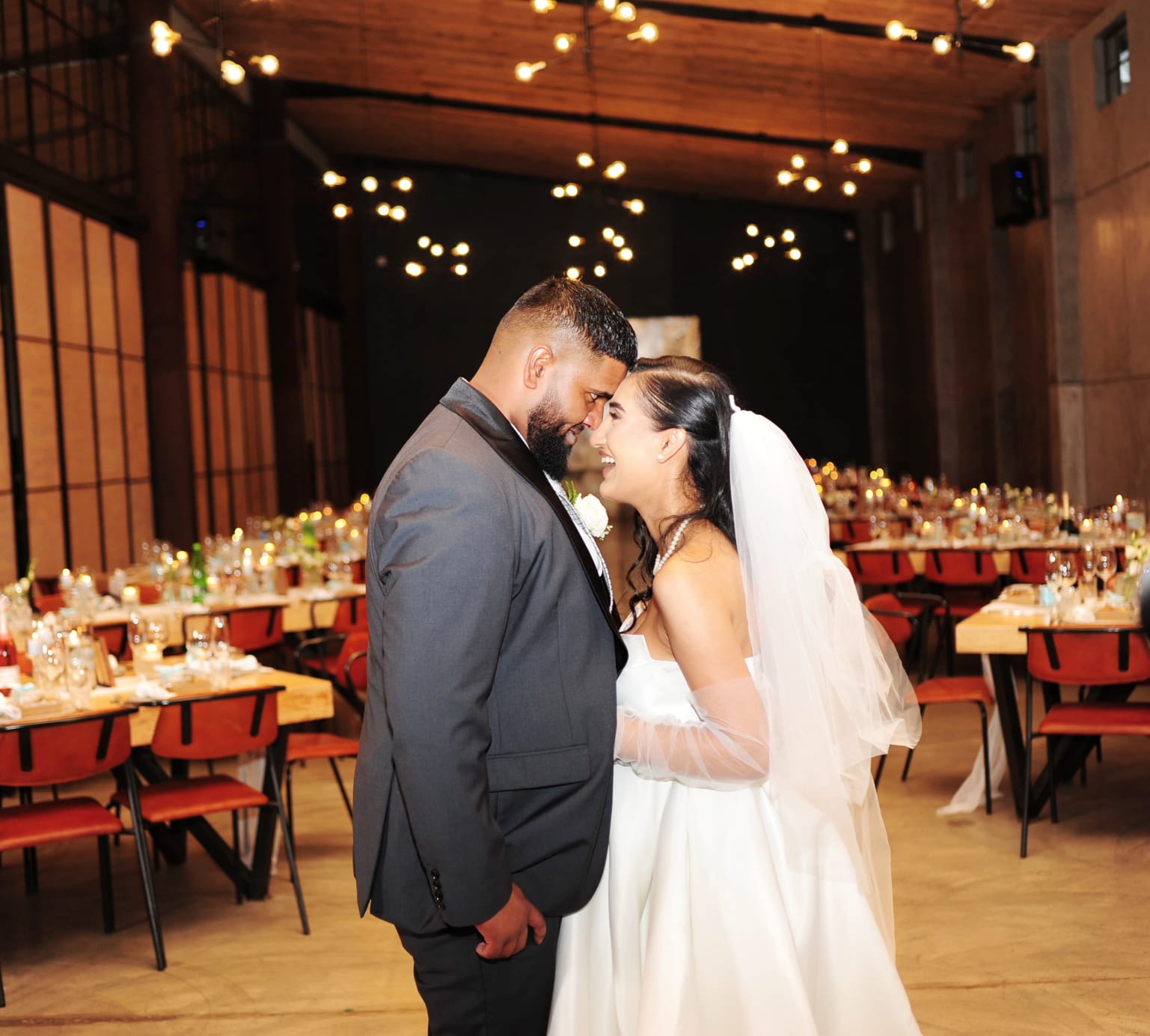 A couple facing each other in a reception hall strung with warm festoon lighting.